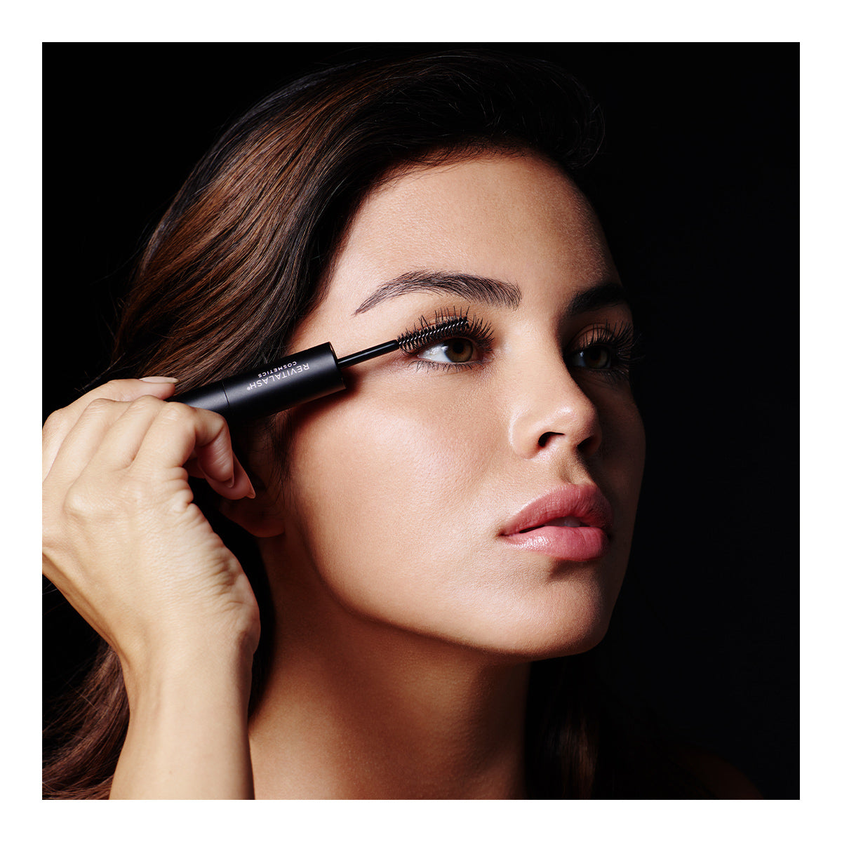 A woman with long, dark hair applies RevitaLashs Double-Ended 2-in-1 Lash Primer & Mascara Volume Set to her upper lashes. She gazes upward against a black background, emphasizing her face and showcasing RevitaLashs ability to enhance natural beauty.