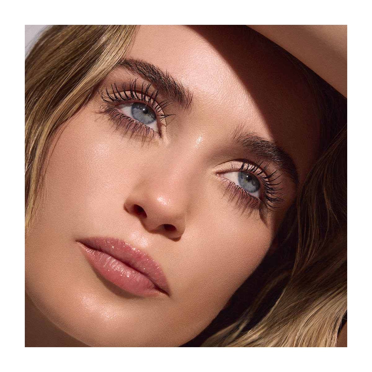 A close-up shows a person with light skin, blue eyes, and long eyelashes appearing 300% longer from using RevitaLash Length Define Tubing Mascara. Their light brown hair frames a shadowed face with a calm expression as they gaze upwards, sunlight enhancing their striking features.