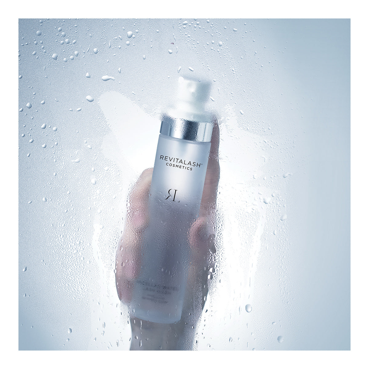 A hand holds a translucent bottle of RevitaLash Micellar Water Lash Wash, Hypoallergenic Makeup Remover (100mL) behind foggy glass. With a metallic cap and minimal branding, this makeup remover is safe for sensitive eyes.