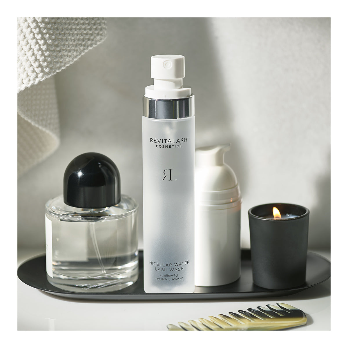 A bathroom counter features RevitaLash Micellar Water Lash Wash, a hypoallergenic makeup remover suited for sensitive eyes, next to a clear perfume bottle, white foaming pump bottle, and lit black candle on a tray, with a white towel and comb nearby.