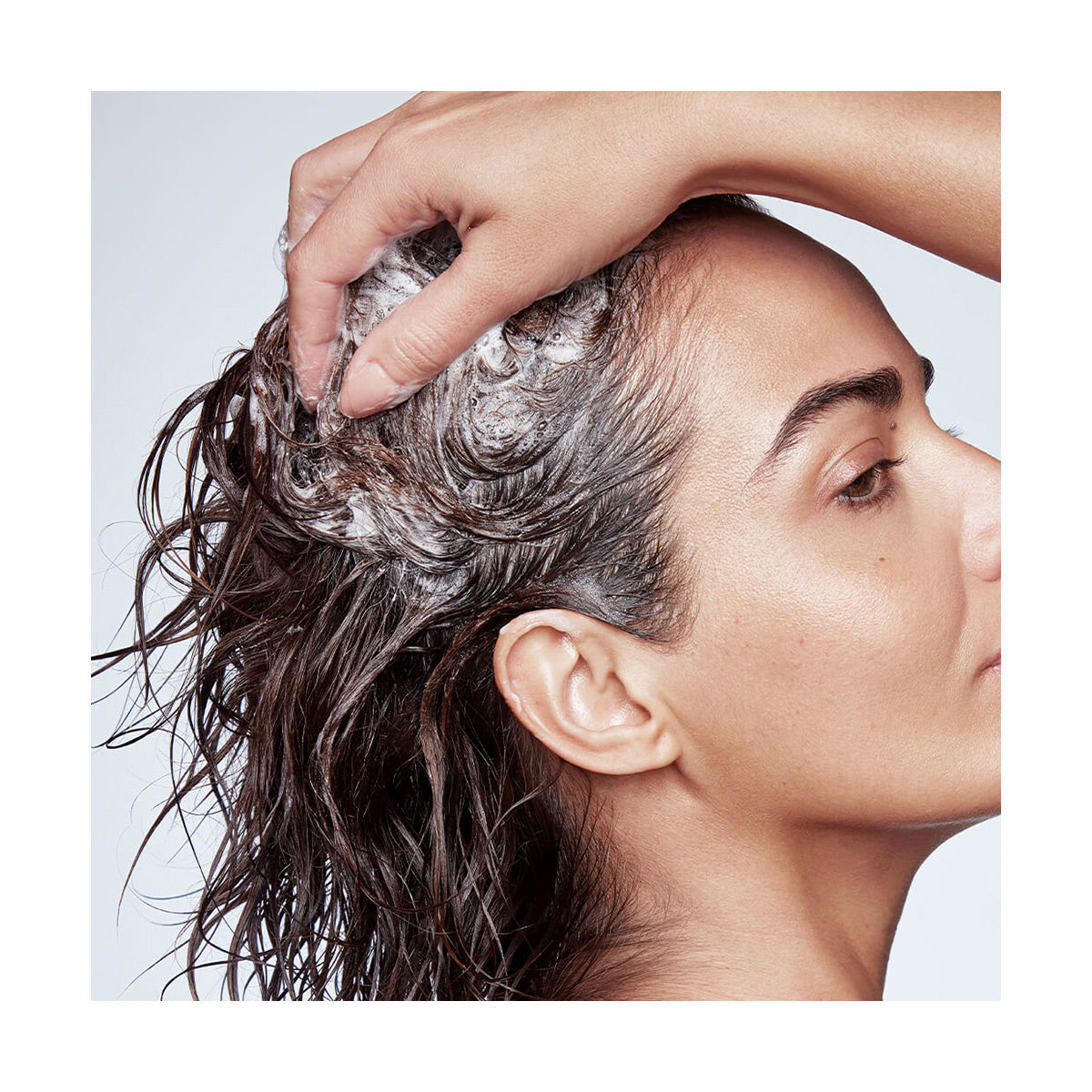 A person massages RevitaLash Thickening Shampoo lather into their scalp using one hand, enhancing optimal scalp health against a light background.