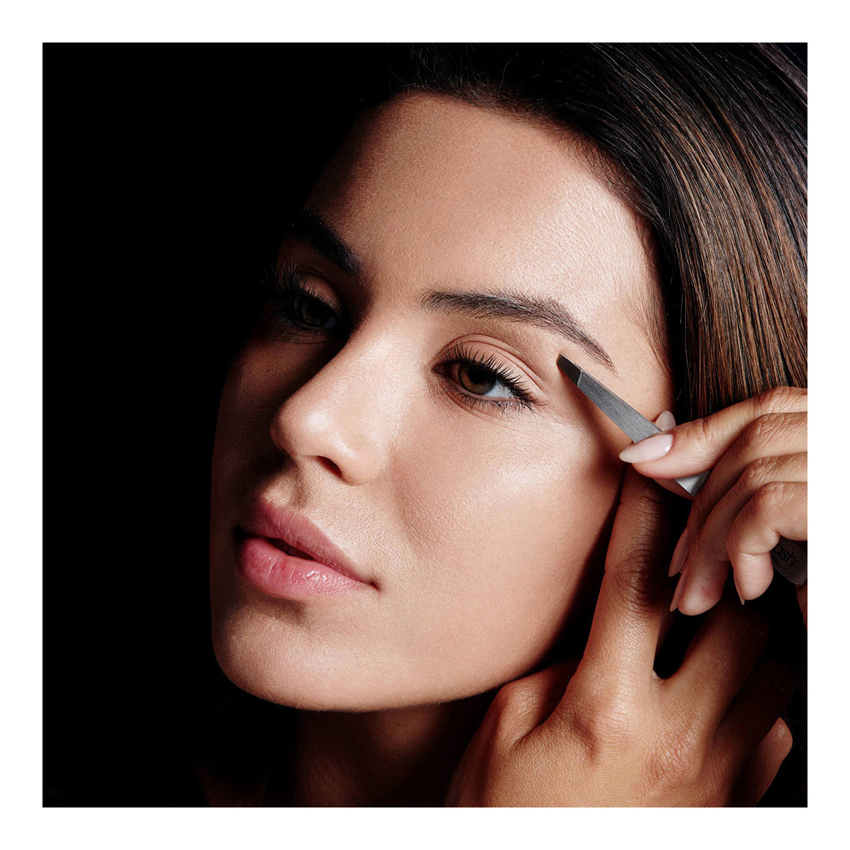 With a focused expression, a dark-haired woman expertly uses the RevitaLash Precision Tweezer, holding it carefully against her face. The scene, featuring a dark background, highlights the meticulous precision and professional-grade care in her routine.