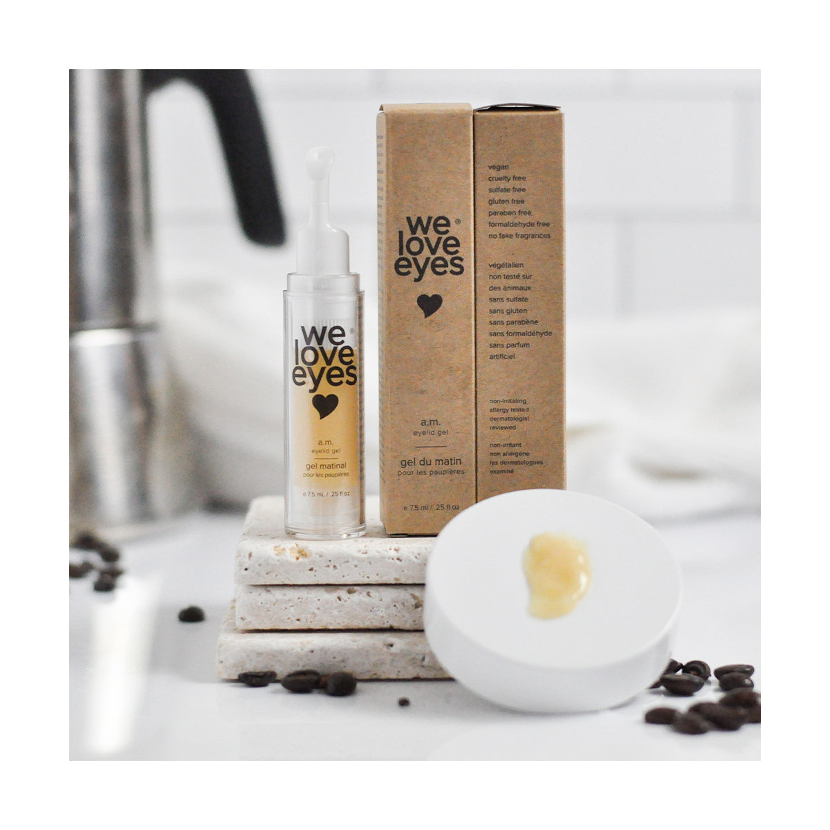 The We Love Eyes - AM Eyelid Gel - 7.5ml bottle is set on stacked white stone coasters with its box nearby. A dab of gel rests on a white lid in the foreground, surrounded by coffee beans suggesting its effectiveness against dark circles, all displayed on a pristine white surface.