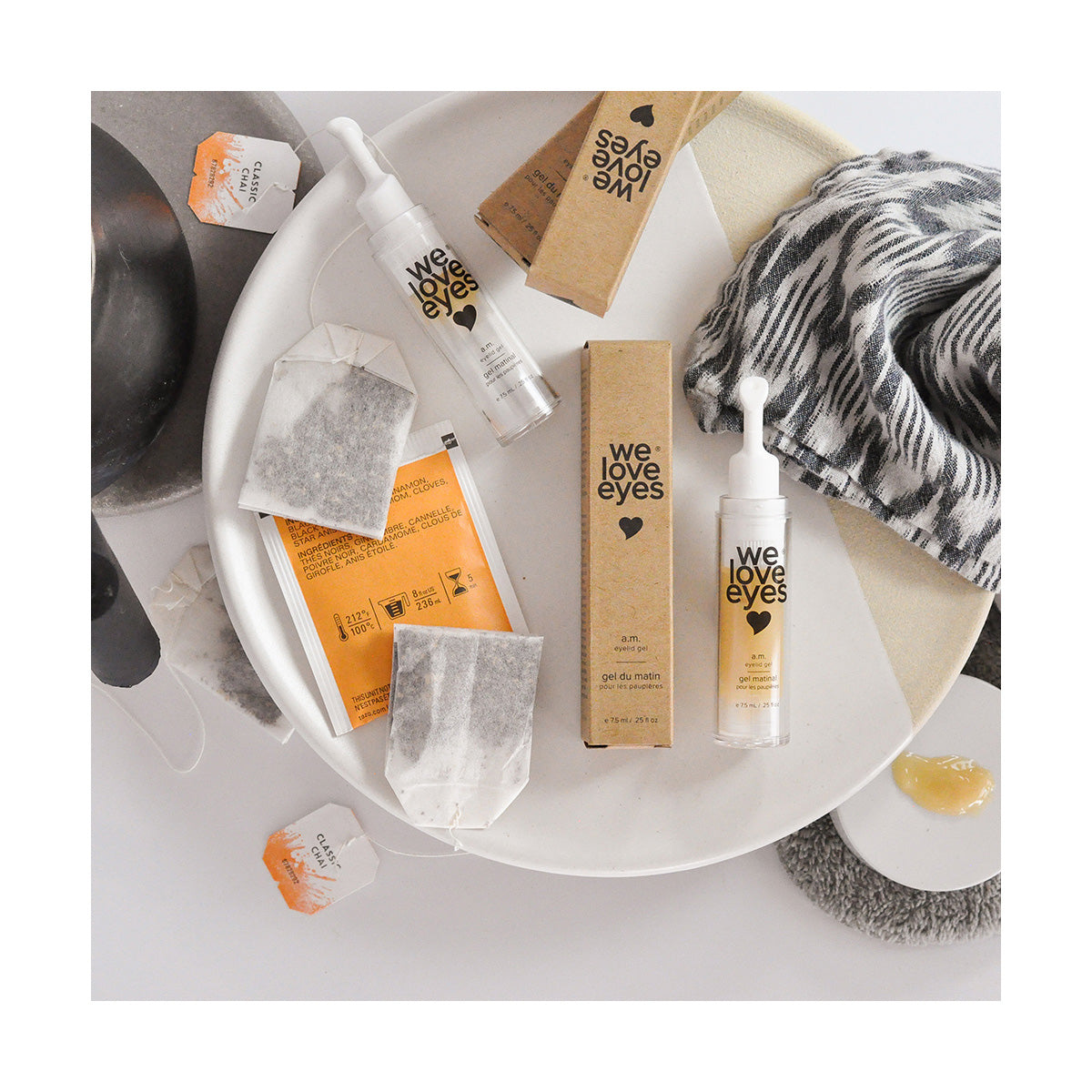 A flat lay showcases We Love Eyes - AM Eyelid Gel and packaging boxes, with a facial brush and tea bags on a white dish to fight dark circles. A gray and white patterned cloth nearby adds to the cozy feel.