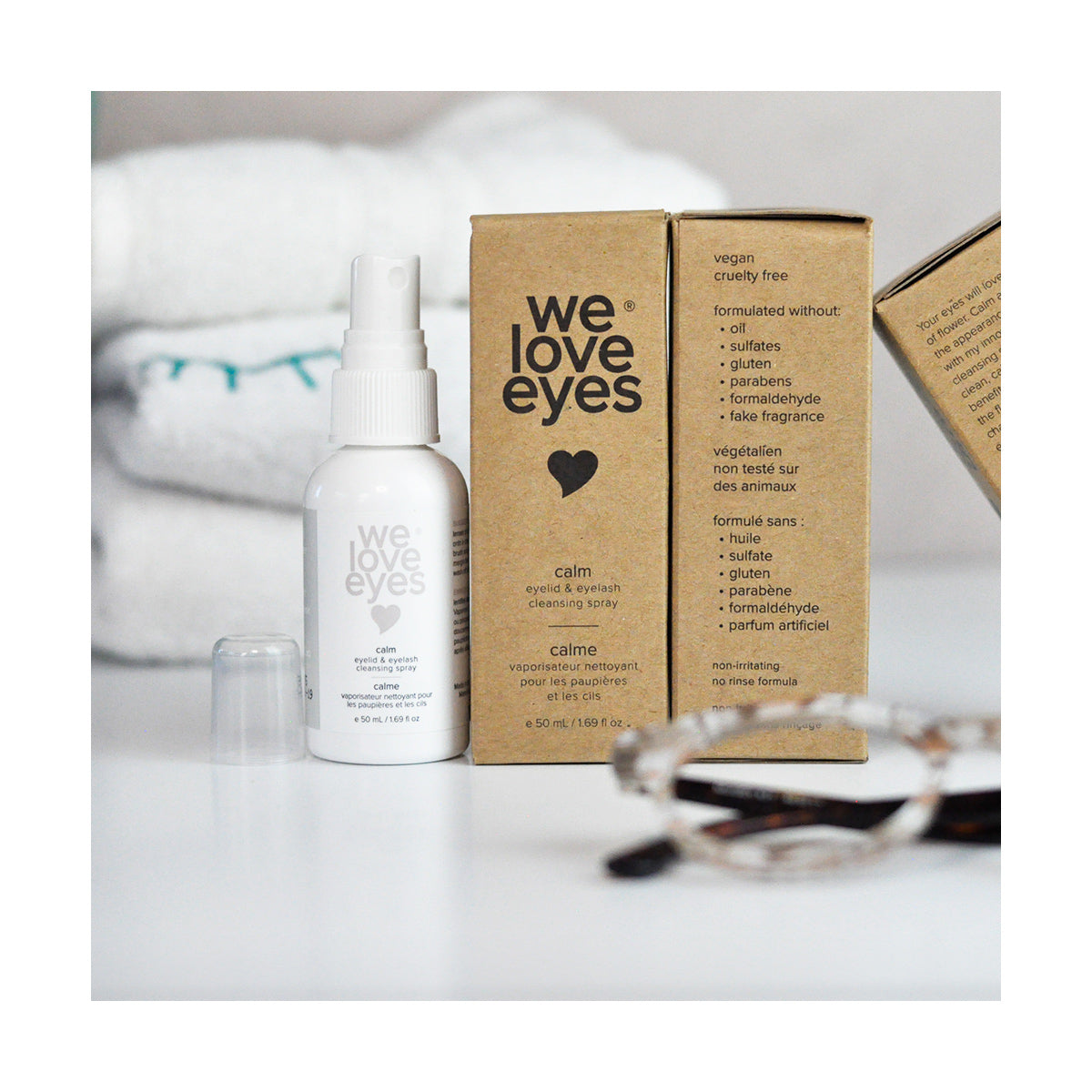 A We Love Eyes Calm Eyelid & Eyelash Cleansing Spray (50mL) is on a white surface with its box. This vegan, cruelty-free solution with HypoChlorous and chamomile excludes harmful chemicals. Glasses and a folded towel blur in the background.