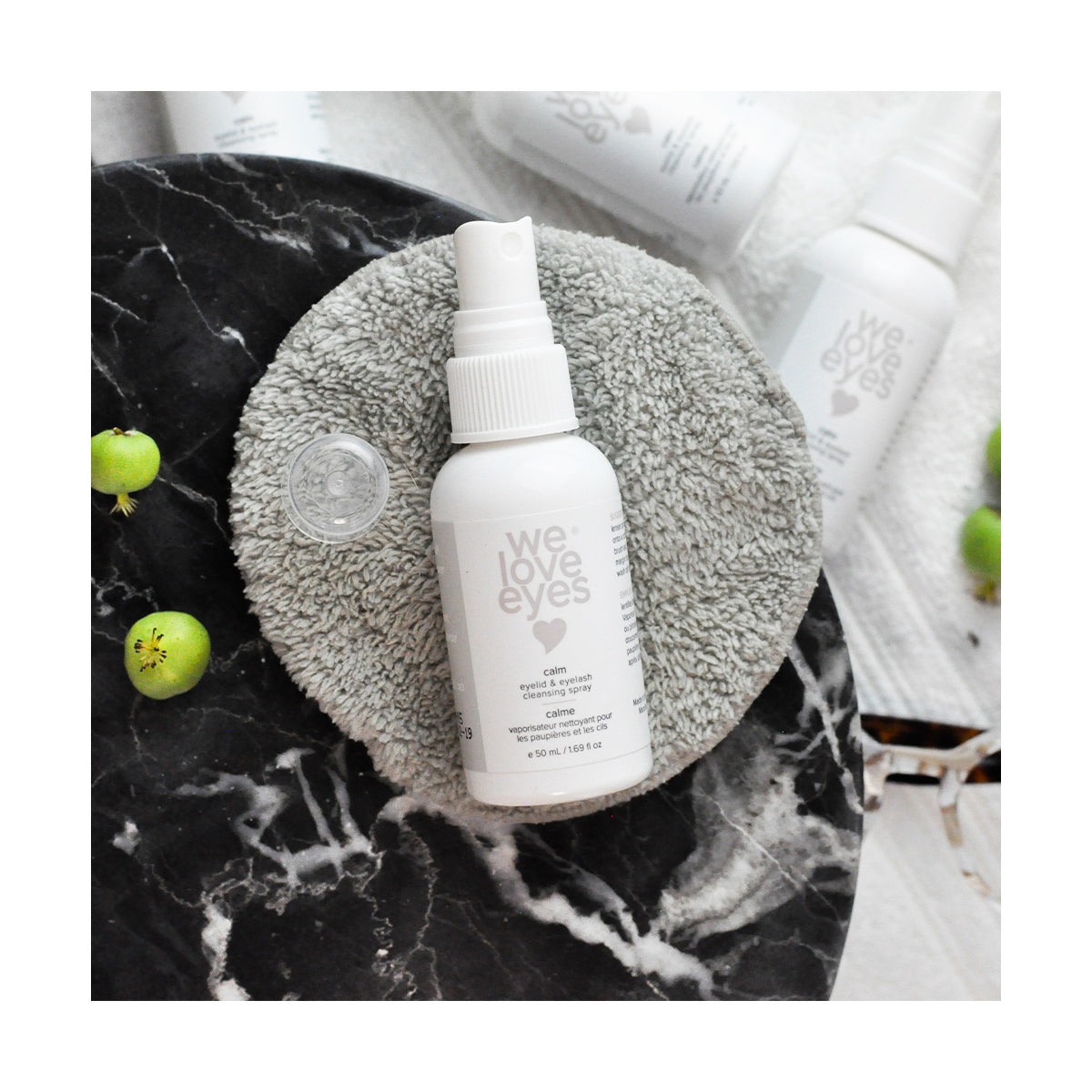 The We Love Eyes Calm Eyelid & Eyelash Cleansing Spray with HypoChlorous, a white 50mL bottle, is placed on a gray pad on dark marble. Nearby are a small clear cap and two green berries, suggesting chamomile tranquility. More bottles form a soft blur in the background.