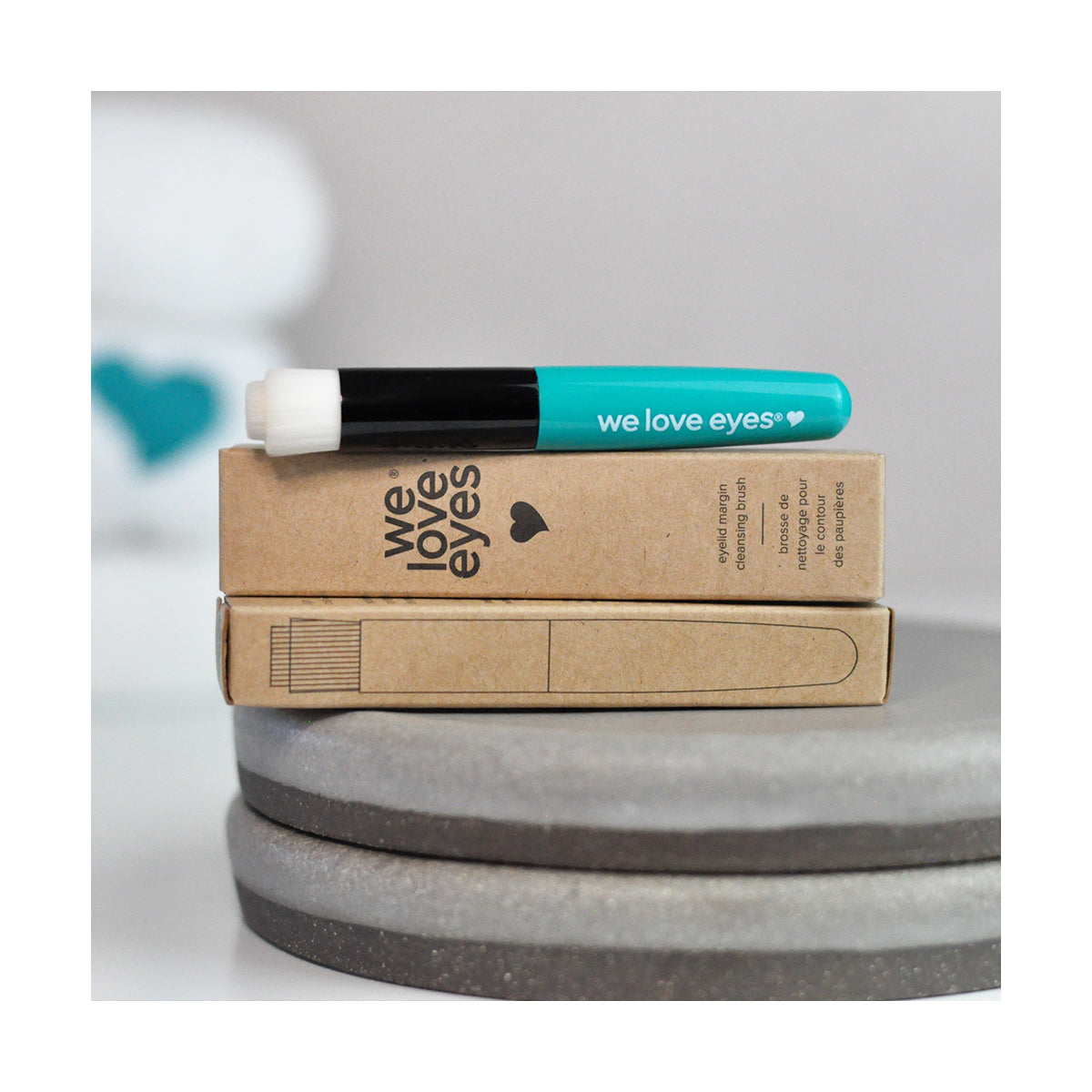 A turquoise We Love Eyes applicator brush for eyelid cleansing is perched on two heart-marked brown boxes. The background softly emphasizes the tiered fiber design crucial for meibomian gland health.