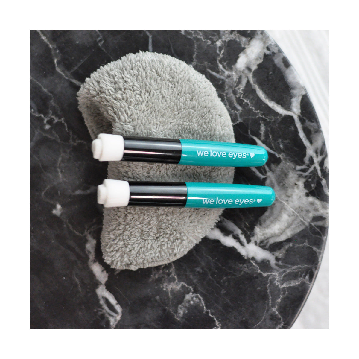 Two teal cylindrical We Love Eyes tubes, labeled as Eyelid Margin Cleansing Brush, sit elegantly on a gray textured pad on a dark marble surface, promoting eyelid hygiene and meibomian gland care.