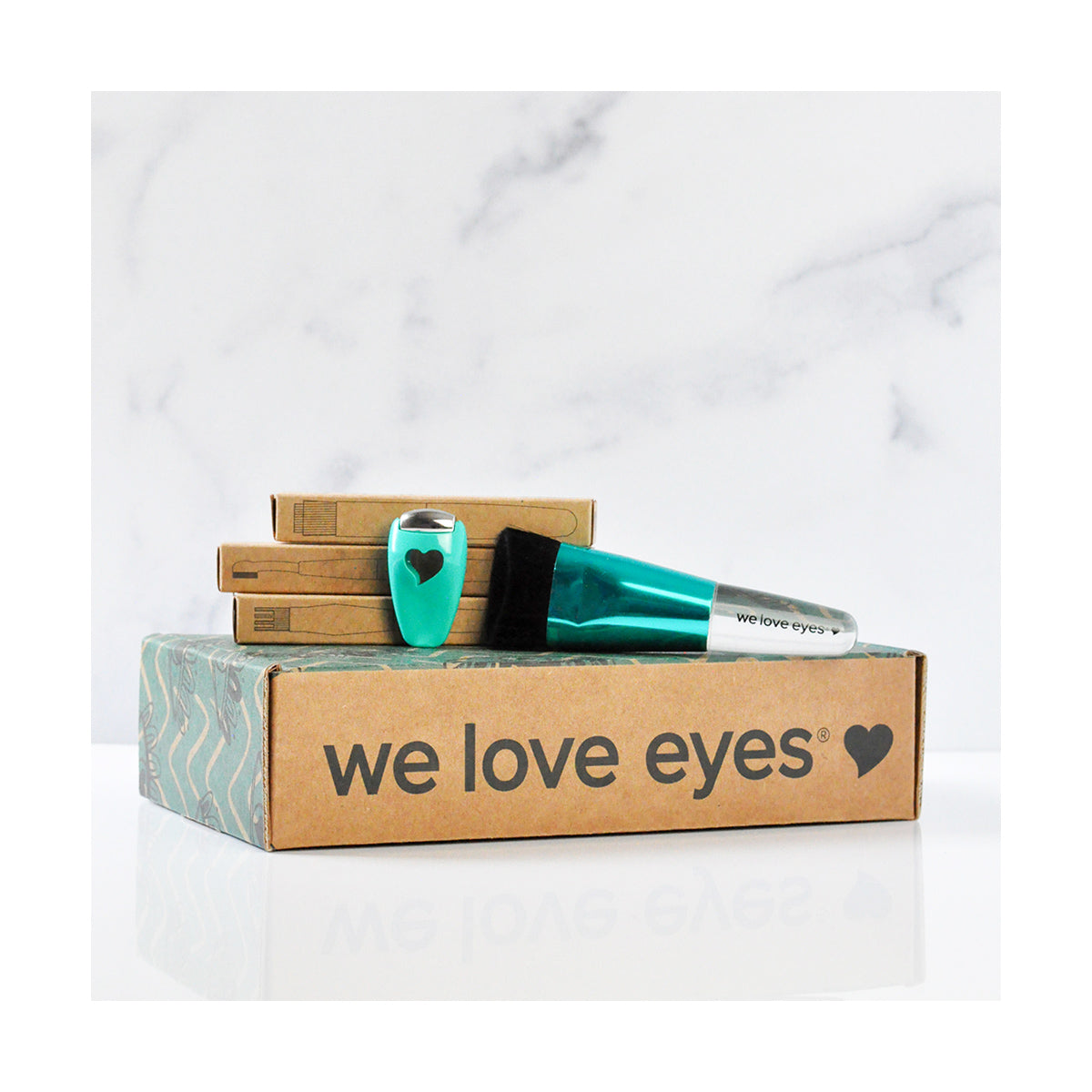 The We Love Eyes Complete Toolkit for Eyelashes, featuring teal bottles and a cleansing brush, is stacked on branded boxes. The minimalist packaging on a reflective white background highlights eyelash follicle health.
