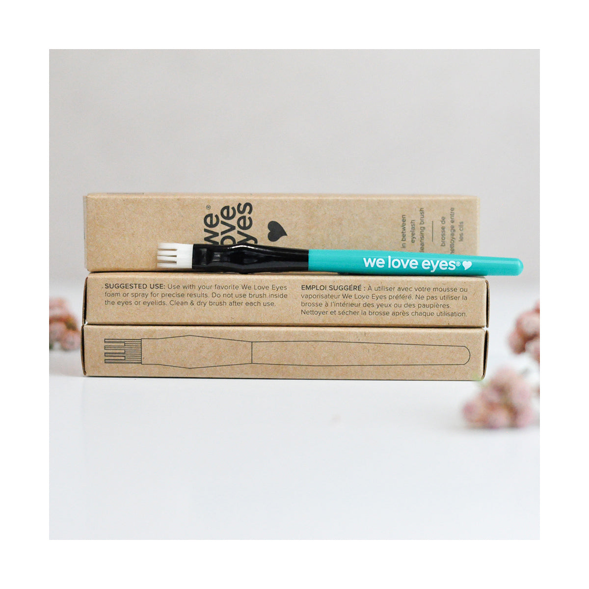The We Love Eyes In Between Eyelash Cleansing Brush, a turquoise scrub shaped like a fork with we love eyes text, rests on two brown boxes that include care instructions. The scene is softened by blurred pink flowers in the background.