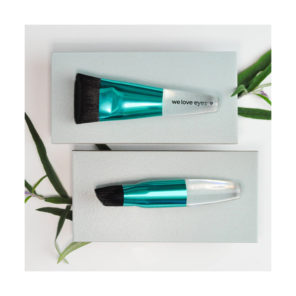 Two makeup brushes with dome-shaped teal handles and black bristles rest on light gray boxes. One is labeled We Love Eyes, highlighting eyelid margin health. Green leaves emphasize the vegan, cruelty-free essence against a white background. Product: Lashfull Thinking brush by We Love Eyes.