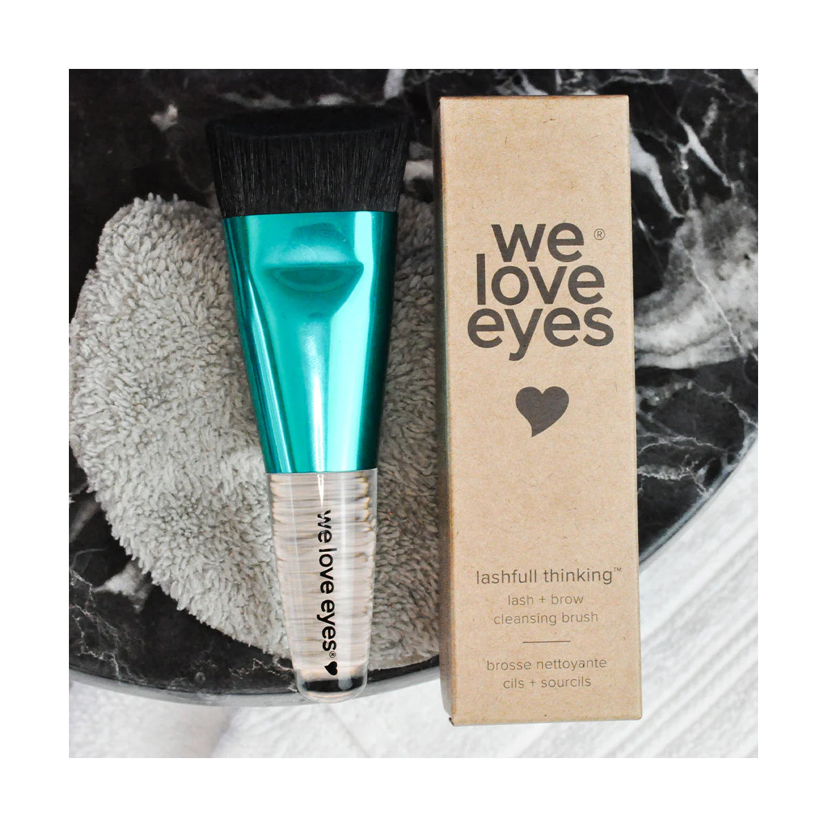 A turquoise, dome-shaped brush labeled We Love Eyes rests beside a brown cardboard box labeled We Love Eyes - Lashfull Thinking lash and brow cleansing brush. Both are on a black marble surface with a gray textured pad underneath, ideal for eyelid margin health.