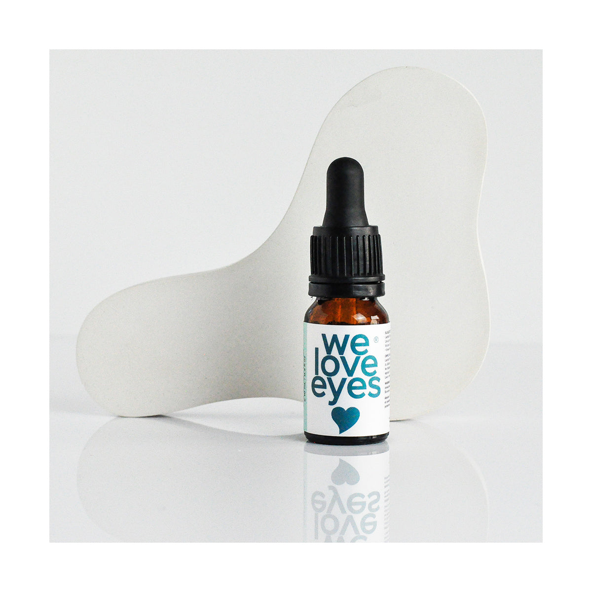 A 10mL We Love Eyes Lashfull Thinkin Lash + Brow Follicle Oil bottle with a dropper cap sits on a reflective surface. The white label reads We Love Eyes in blue, suggesting lash and brow benefits. An abstract white shape in the background adds elegance to the scene.