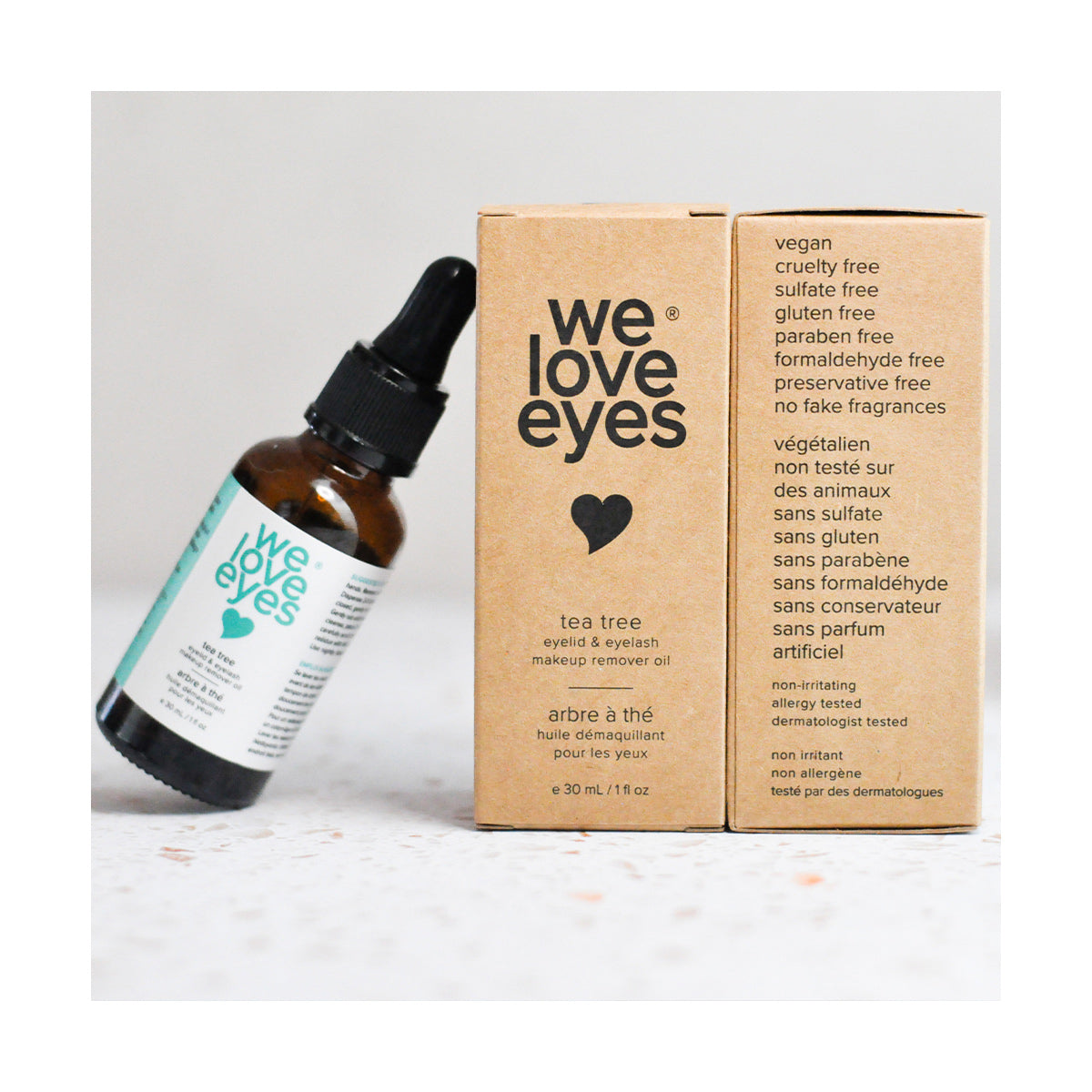 A We Love Eyes 30mL bottle of 100% All Natural Tea Tree Makeup Remover Oil, designed to remove waterproof makeup, leans left. Two boxes emphasize attributes like vegan, cruelty-free, and sulfate-free. Packaging features a heart icon and product specifics.