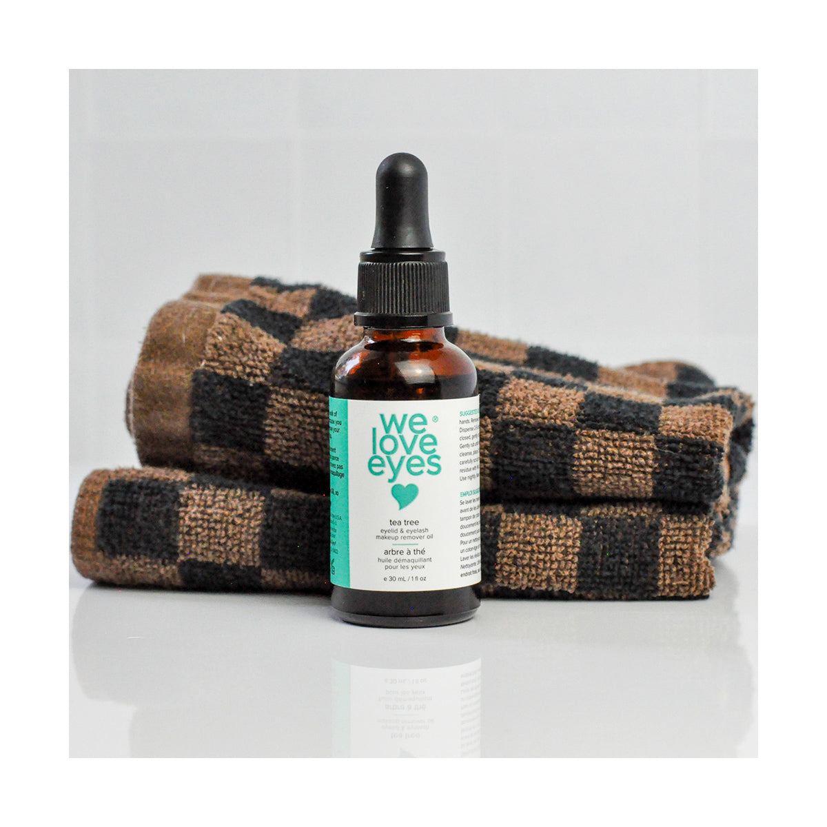 A bottle labeled We Love Eyes - 100% All Natural Tea Tree Makeup Remover Oil for removing waterproof makeup and eyeliner sits in front of folded brown and black checkered towels. The plain white background highlights its simple elegance.