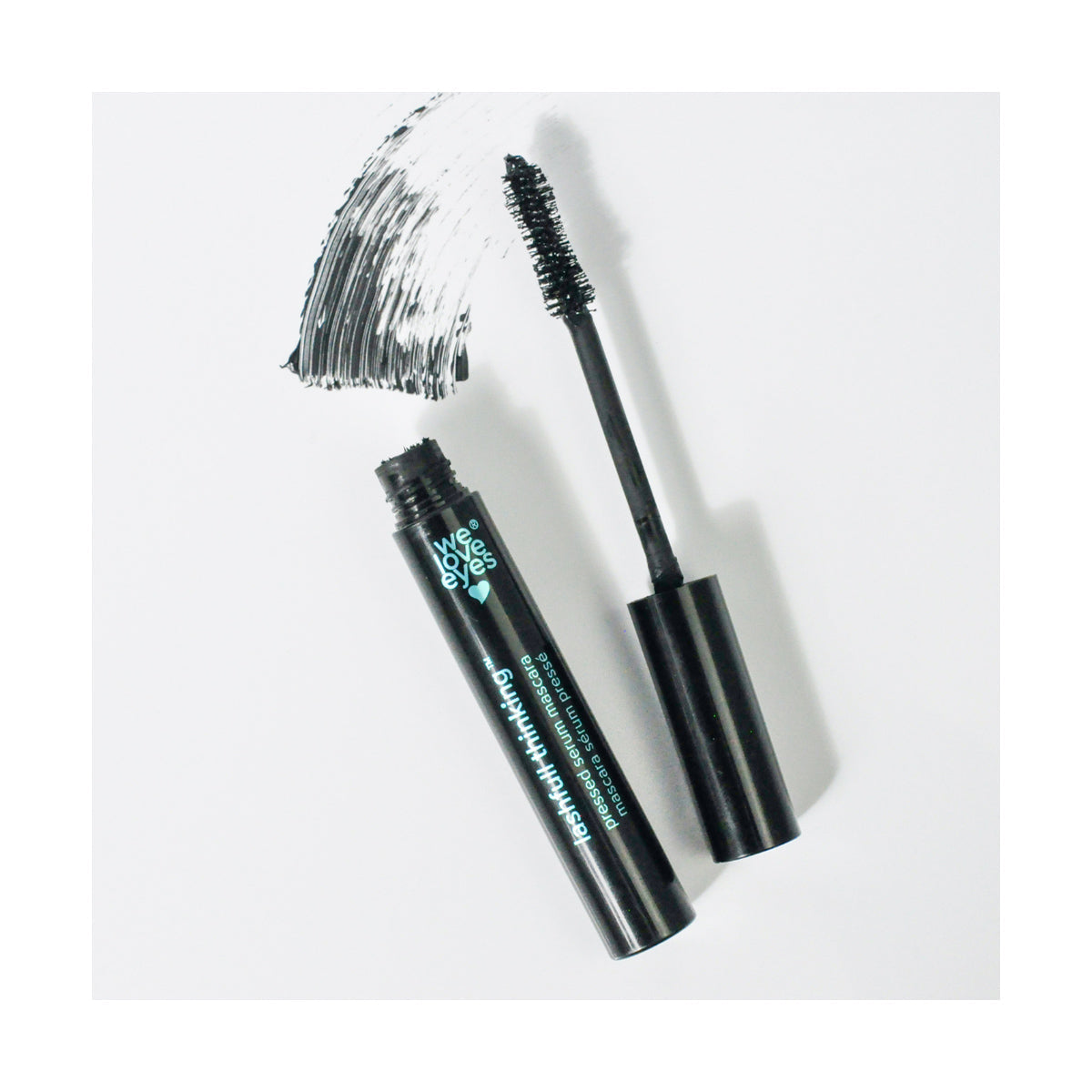 A tube of We Love Eyes Lashfull Thinking BLACK Pressed Serum Mascara with Widelash lies on a white surface. The wand, coated in black mascara, creates a brush stroke above the tube, while the sleek packaging text is partially visible.