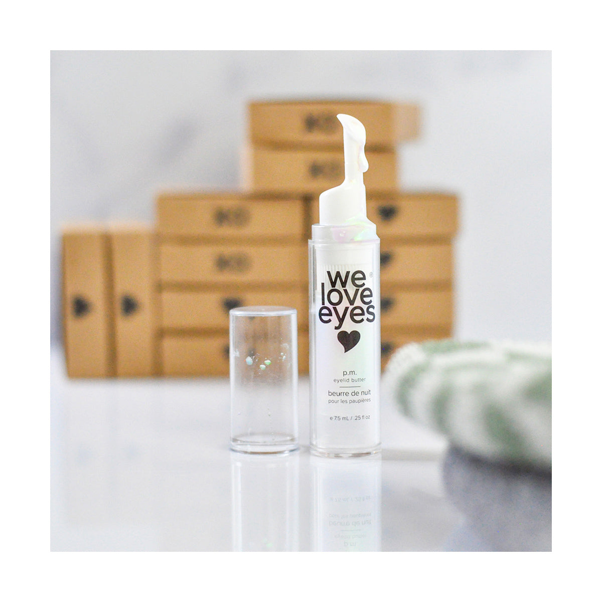 An open bottle of We Love Eyes - P.M. Eyelid Butter - 7.5ml sits with its cap beside it against a backdrop of heart-patterned cardboard boxes. Designed for delicate eyelid skin, a soft cloth peeks out on the right.