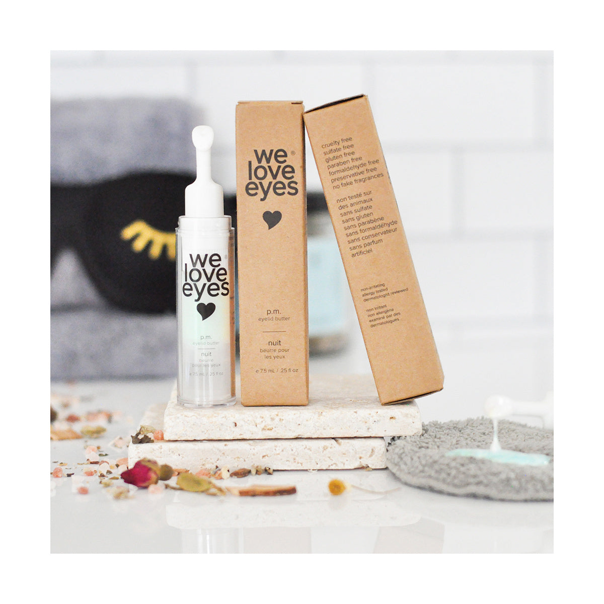 A glass dropper bottle of We Love Eyes - P.M. Eyelid Butter - 7.5ml rests on a white marble block next to its brown box, designed to support eyelid skin and reduce water loss, with a gray washcloth, black sleep mask, and dried flowers scattered on a white surface in the background.