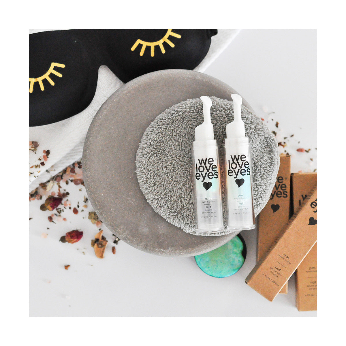 The arrangement includes two We Love Eyes - P.M. Eyelid Butter bottles on a gray pad, alongside a black sleep mask with eyelashes design, dried flowers, and product boxes. This cozy setup offers essentials for protecting the eyelid skin barrier against transepidermal water loss.