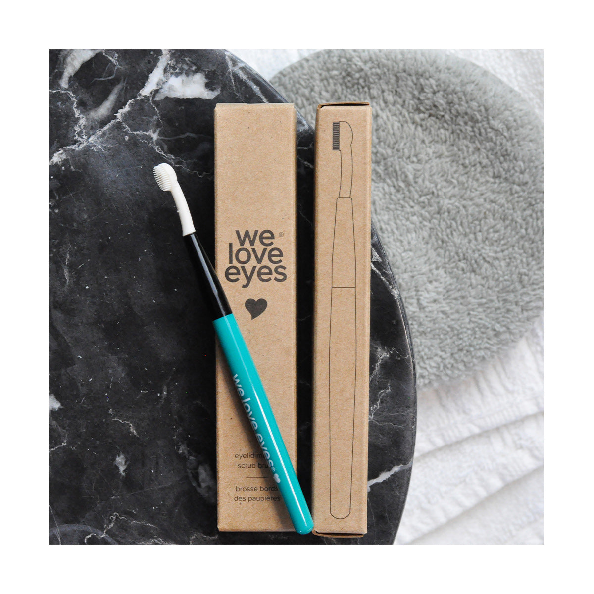 The We Love Eyes Eyelid Margin Scrub Brush, with a turquoise handle, sits next to its we love eyes brown cardboard packaging on a dark marble surface. Paired with a gray cloth and white towel, its perfect for cleansing eyelid margins and tackling issues like demodex collarettes.
