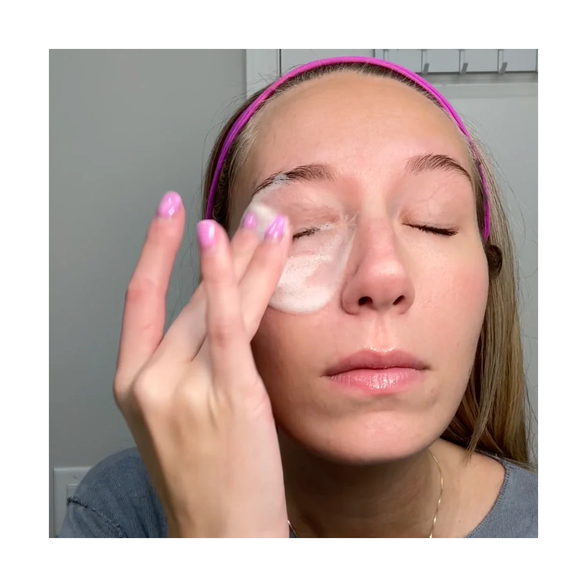 In a neutral-toned setting, a person with a pink headband and light nail polish gently applies We Love Eyes SuperGLA eyelid & eyelash foaming cleanser, rich in omega-6 fatty acid, to their closed eye using a soft pad as part of their skincare routine.