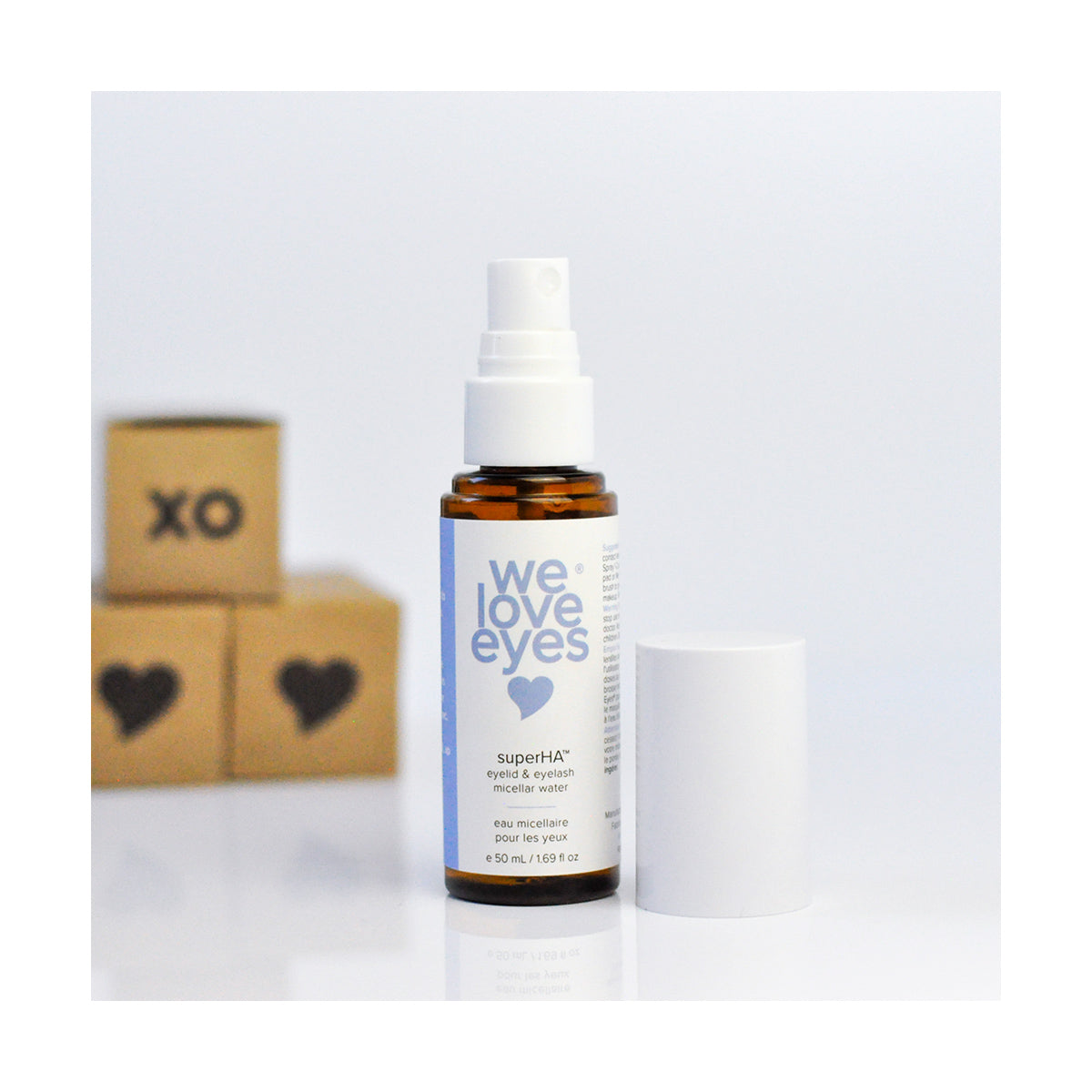 A bottle of We Love Eyes superHA Eyelid & Eyelash Micellar Water Make-up Remover (50mL) rests on a reflective surface. In the background, three brown cubes are visible; one displays XO and another has a heart symbol.