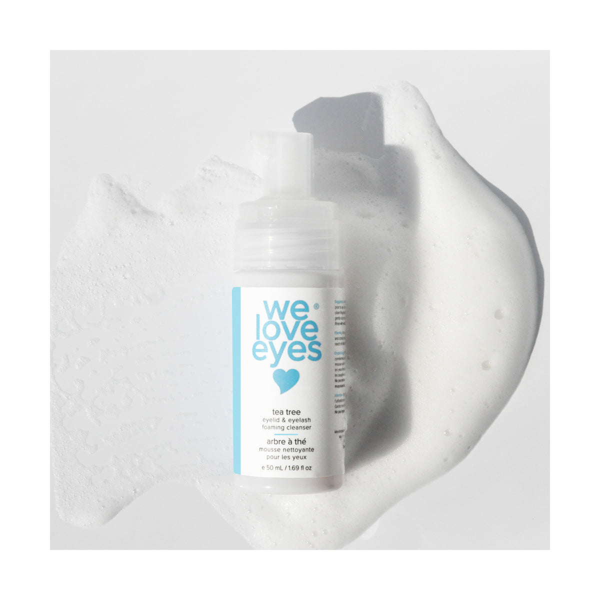 The We Love Eyes: Tea Tree Eyelid Foaming Cleanser rests on a pristine white surface surrounded by bubbles, featuring a pump top and light blue label. Its infused with Australian Tea Tree Oil to support eyelid margin health.