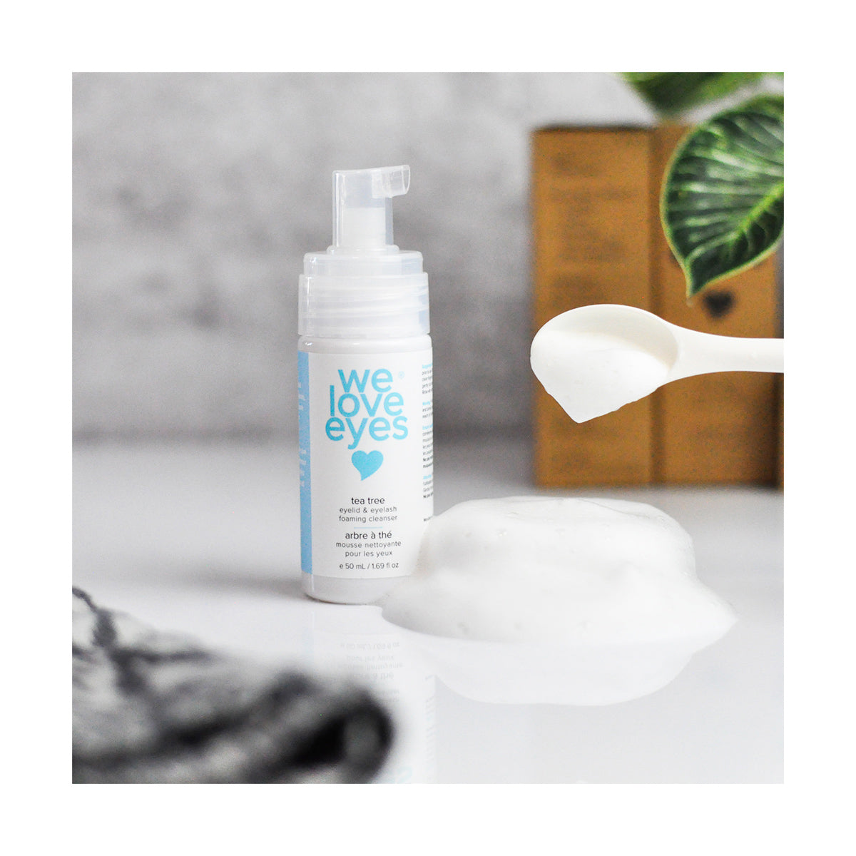 A white bottle of We Love Eyes: Tea Tree Eyelid Foaming Cleanser with blue labeling stands upright, dispensing foam onto a white surface. A spoon holds more foam, ensuring eyelid margin health with Australian Tea Tree Oil. A plant and wooden box are visible in the background.
