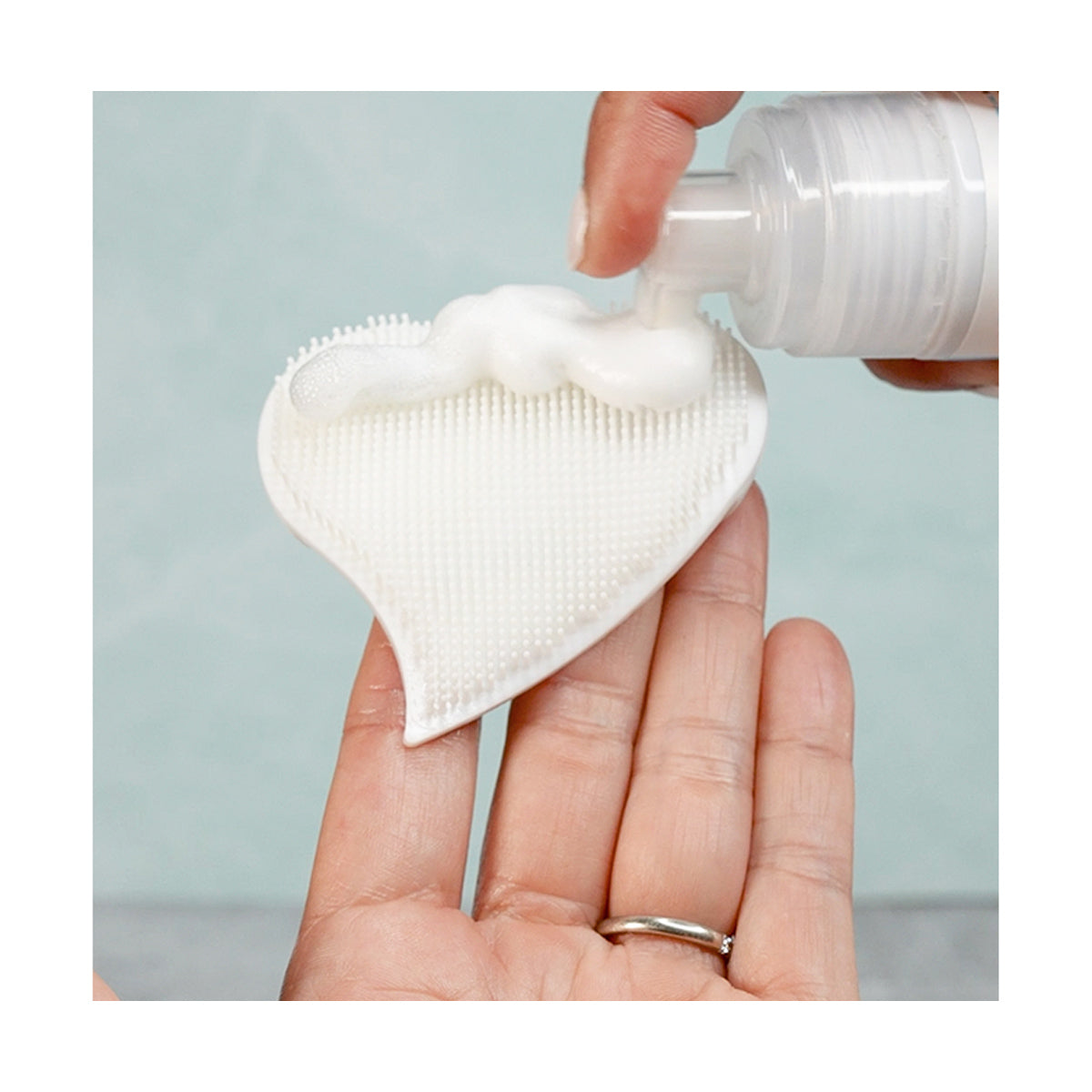 A persons hand holds a white, heart-shaped silicone scrub pad while another dispenses We Love Eyes Tea Tree Eyelid & Eyelash Foaming Cleanser from a white pump bottle onto the pad. The background is a soft, blurred teal color.
