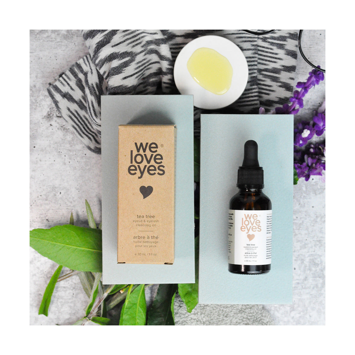 A box and the We Love Eyes - All Natural Tea Tree Eyelid Cleansing Oil - 30ml bottle are set on a textured surface. Nearby, a small dish with oil, purple flowers, and leafy greens enhances the aromatic setting while supporting healthy meibomian oil glands.