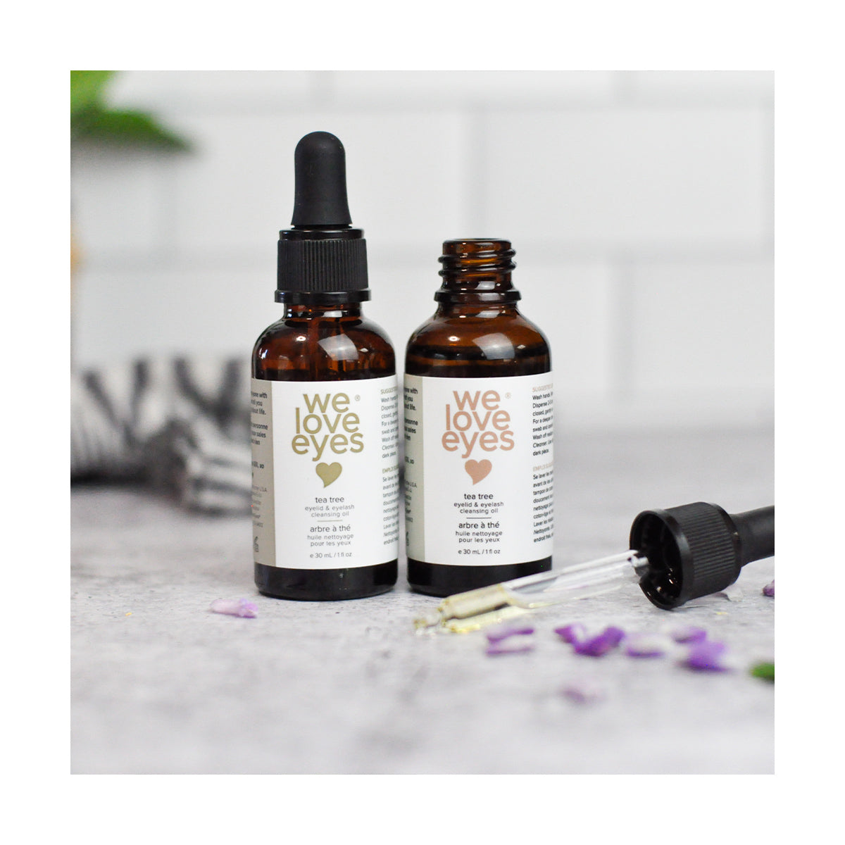 Two brown glass bottles of We Love Eyes- All Natural Tea Tree Eyelid Cleansing Oil - 30ml, featuring droppers—one open, one closed—rest on a gray surface with purple petals. A blurred striped fabric is in the background, highlighting these Australian Tea Tree Oil-infused natural eye care products.