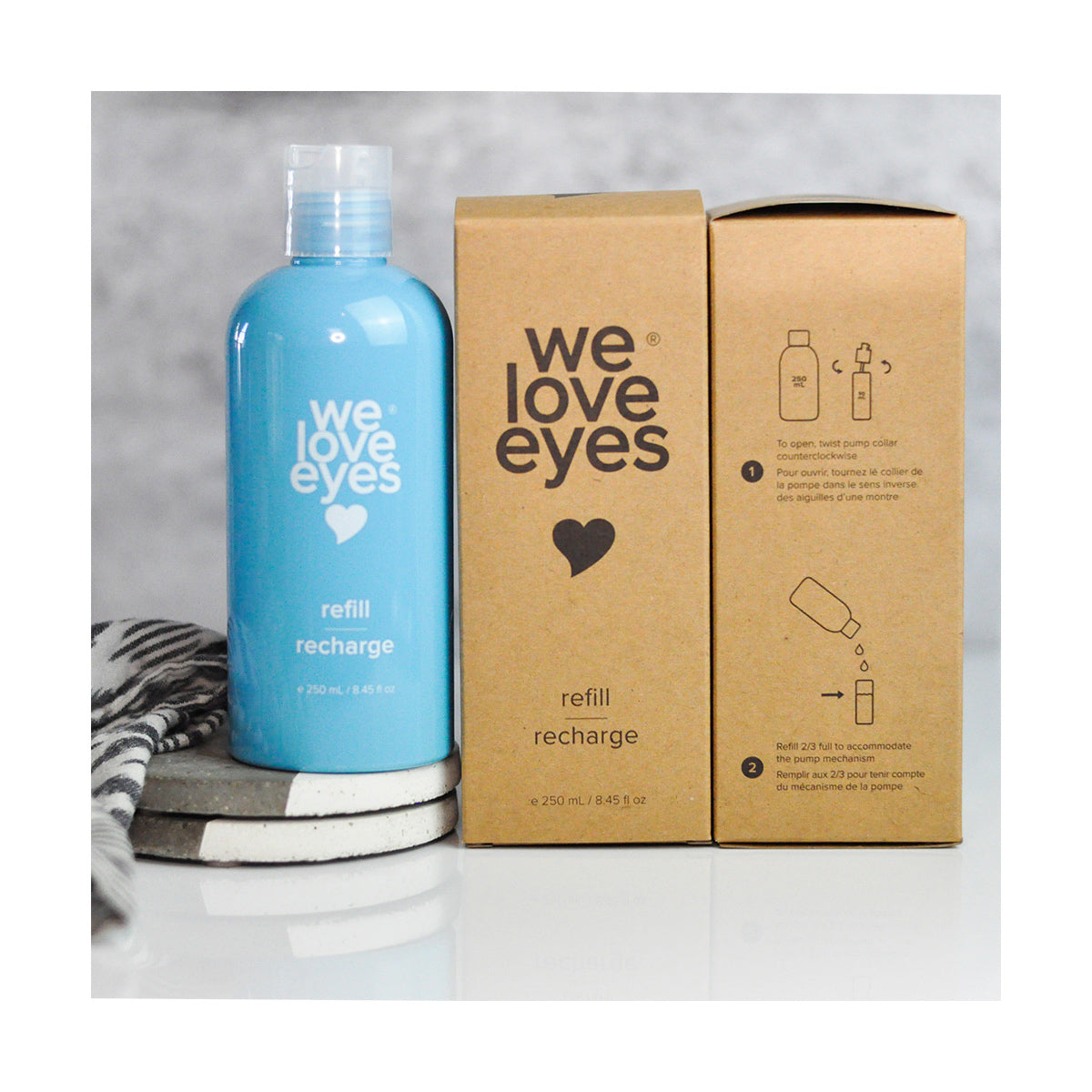 A We Love Eyes Tea Tree Eyelid Foaming Cleanser Refill in a blue bottle and two brown cardboard boxes, both featuring a heart and crafted from post-consumer recycled plastic, sit alongside a striped cloth and round coasters. Recycling instructions are included.