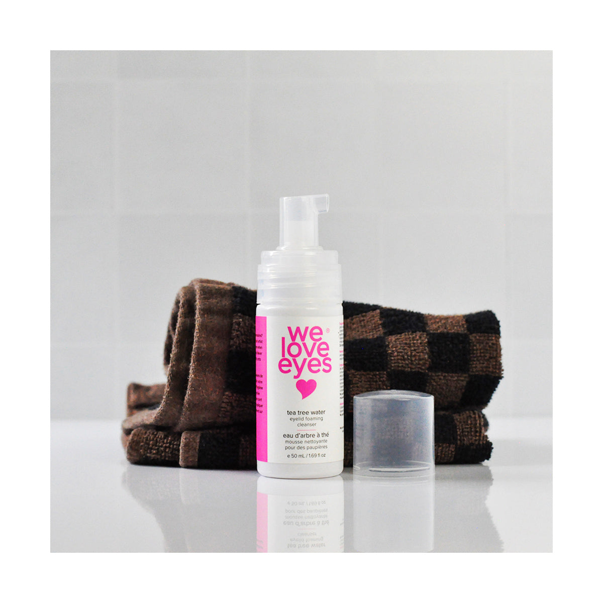 The We Love Eyes Tea Tree Water Eyelid Foaming Cleanser, in a clear bottle with a white and pink label, rests on a reflective surface next to its cap and a folded dark brown and black checkered towel, highlighting its role as a refreshing pH-balanced cleansing spray.