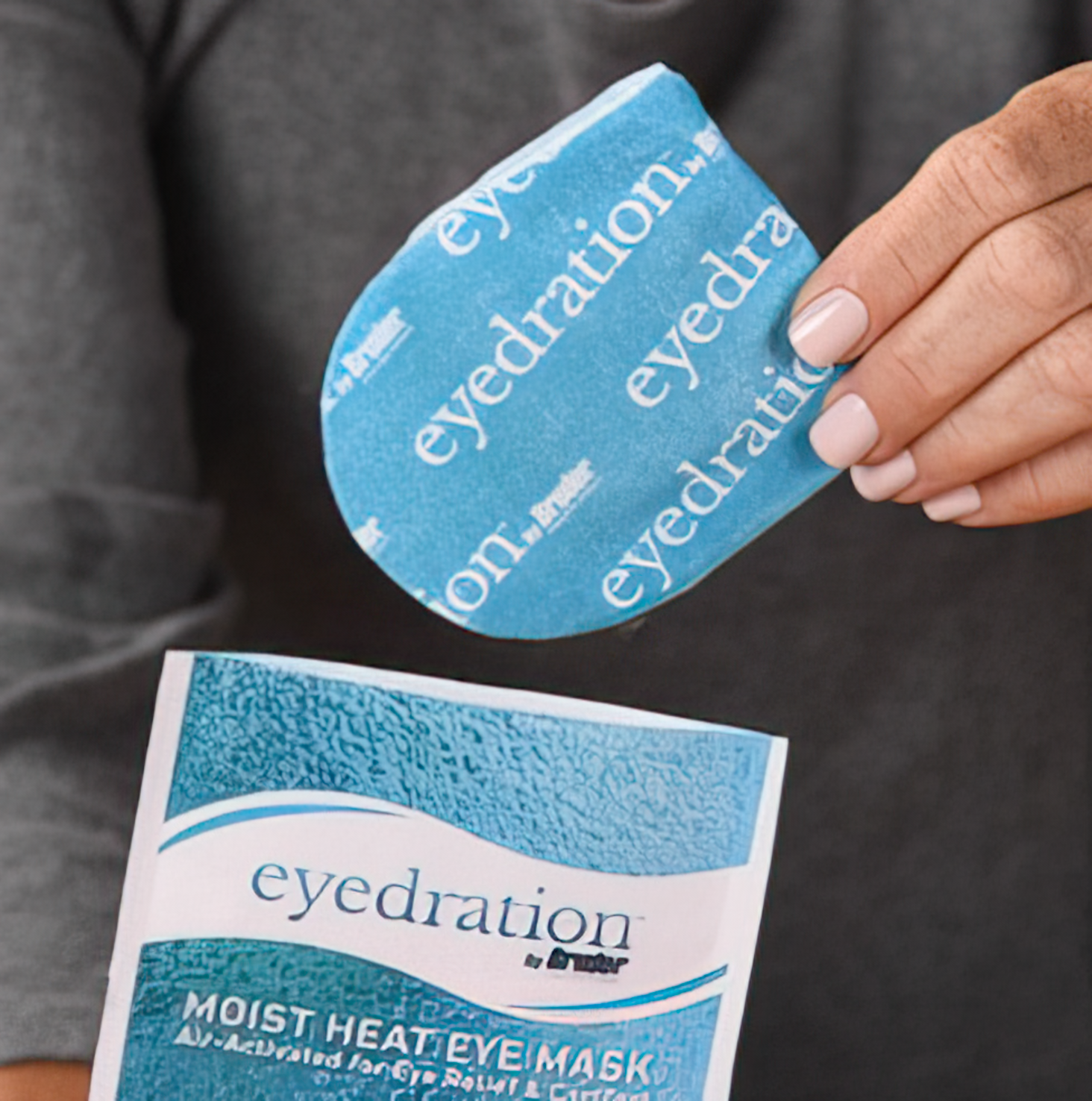A person holds an eyepad labeled eyedration over a partially opened packet. The packet is also labeled eyedration Moist Heat Eye Mask and features a blue and white design. The person is wearing a gray garment.