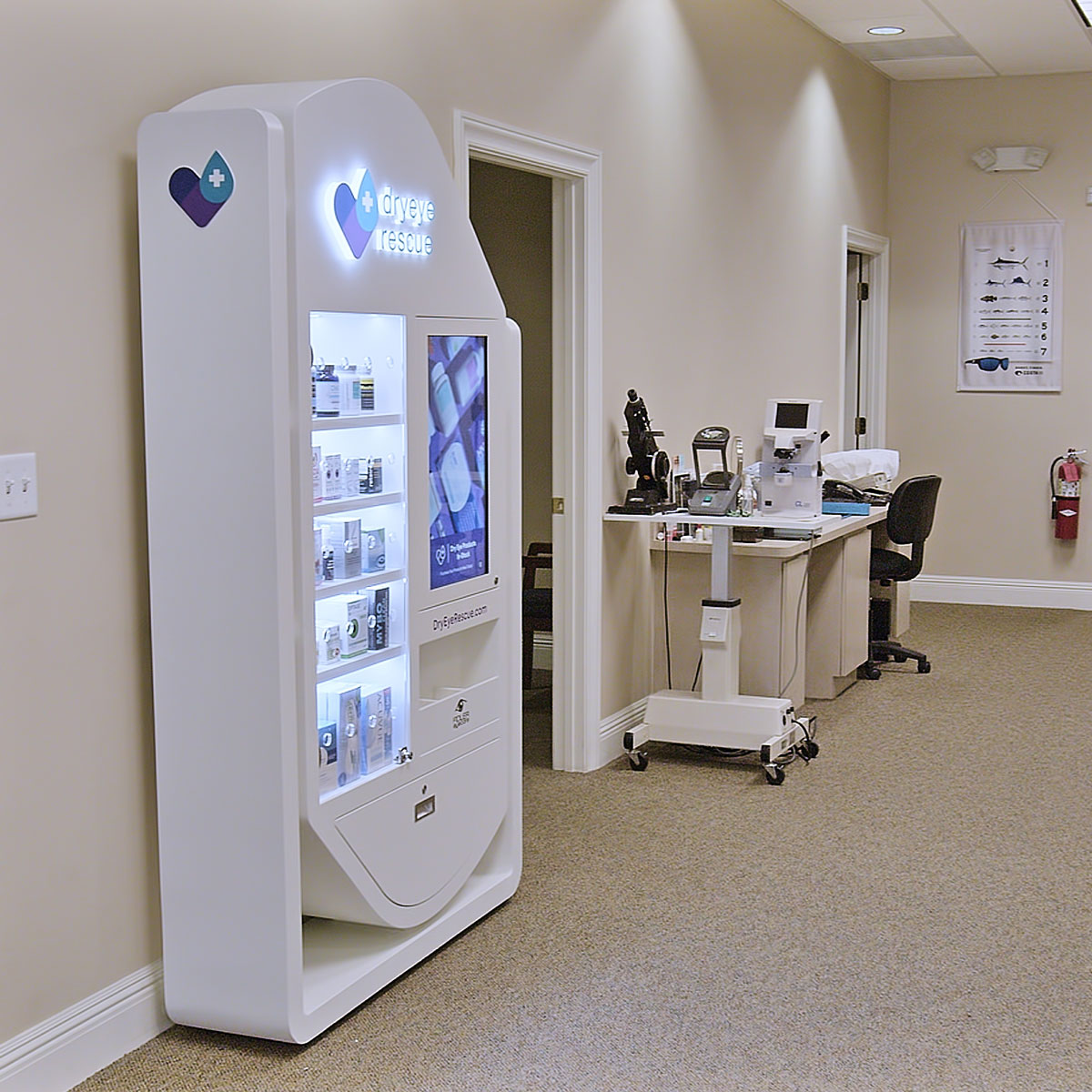 A white DryEye Rescue Standing Floor Display with wheels, stocked with health products, stands near a TV in a medical office corridor. Located by an eye chart and chairs, it helps alleviate patients dry eye concerns.