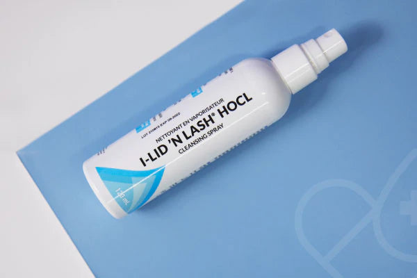 A white bottle labeled I-LID N LASH HOCL Cleansing Spray with blue and silver design rests on a blue surface. The cap is white, and the blue background features a subtle heart and cross symbol.