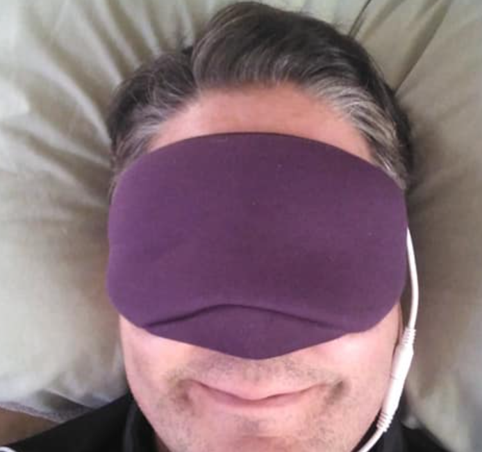 A person with dark hair lies serenely against a pillow, smiling slightly while wearing earbuds and a purple Moisture Eyes Heavenly Heat Pad with Infrared Technology by Dry Eye Guys. The USB-powered mask provides soothing warmth from microwave clay beads.