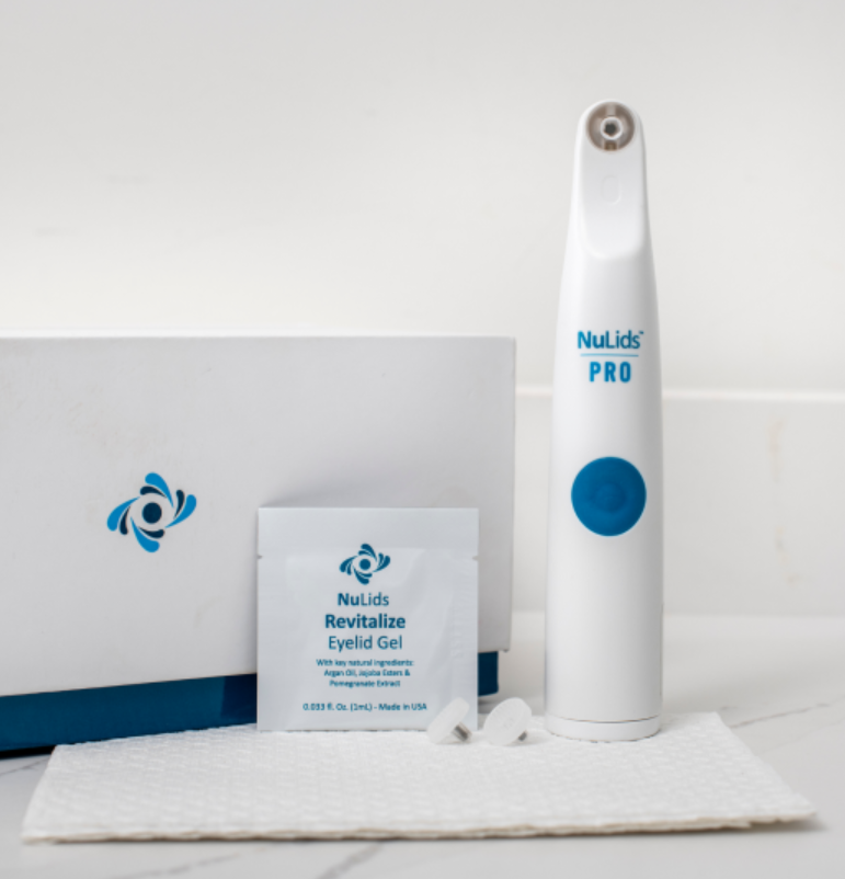 A NuLids Pro In-Office Eyelid Cleaner by NuSight Medical stands upright next to a box and packet labeled NuLids Revitalize Eyelid Gel, highlighting its role in advanced eyelid treatment. The scene includes small pads for dry eye care, set on a white mat against a neutral background.