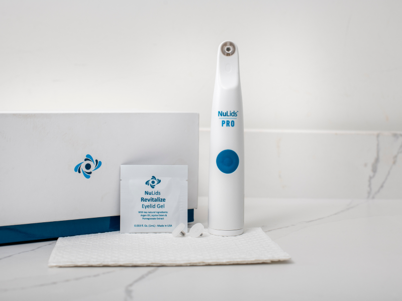 A white NuLids Pro In-Office Eyelid Cleaner from NuSight Medical stands upright on a counter, featuring a blue button for effective eyelid treatment. Beside it are NuLids Revitalize Eyelid Gel in a sachet and a textured cloth. A box with the brands logo sits nearby, emphasizing its role in dry eye care.