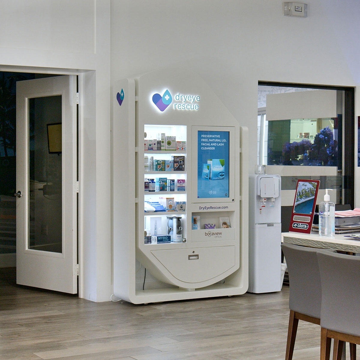 A clean, modern room features a white DryEye Rescue vending machine stocked with eye care products. Next to it, the PKG: Hybrid Standing Floor Display offers informational pamphlets. A water dispenser is nearby, and a fish tank shimmers in the background.