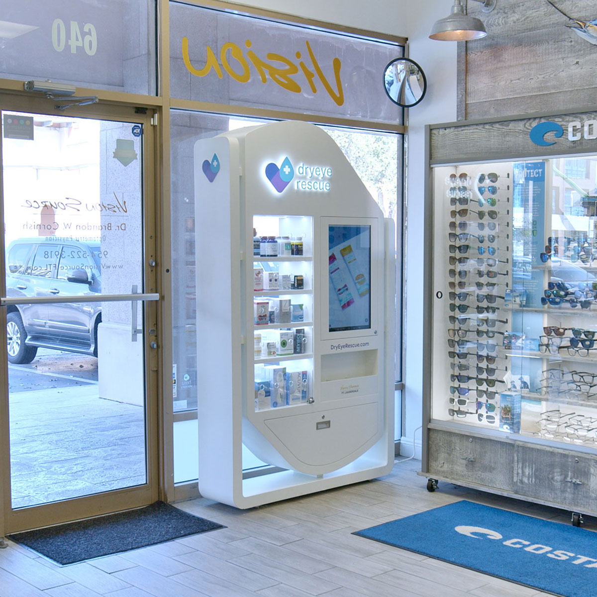 A modern eyewear store interior includes a DryEye Rescue vending machine next to the PKG: Hybrid Standing Floor Display of eyeglasses. A glass door labeled “Vision” leads outside, and a blue mat says “Costco.” Nearby, a TV engages customers with dry eye content.