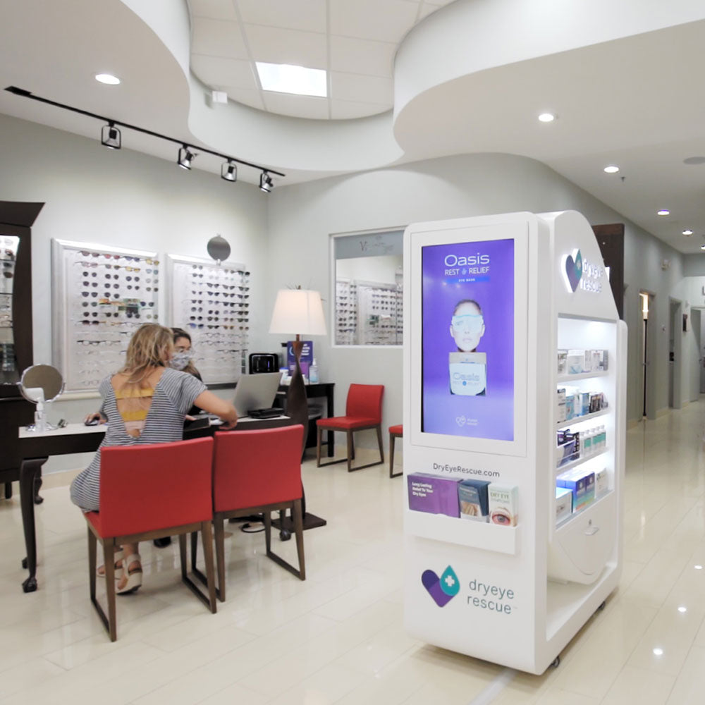 A contemporary optical store boasts a prominent DryEye Rescue PKG: Large Standing Floor Display Free Rent (Large) on the right. A couple chats with an eye specialist in the background, while shelves of eyeglasses line walls beneath ceiling circular lights.