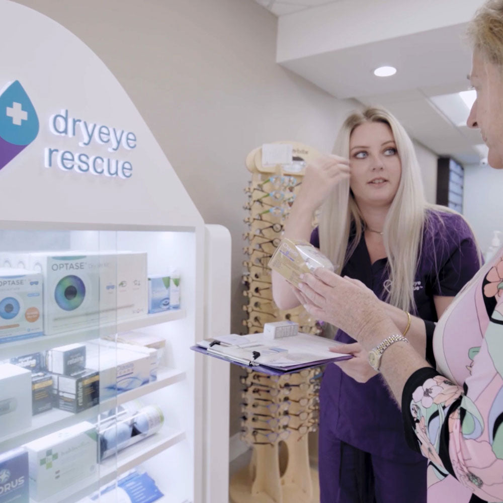 A woman in a purple uniform advises another with a box at the DryEye Rescue eye rescue display featuring various products. Nearby, an impressive PKG: Large Standing Floor Display Free Rent showcases eyeglasses in the well-lit room.