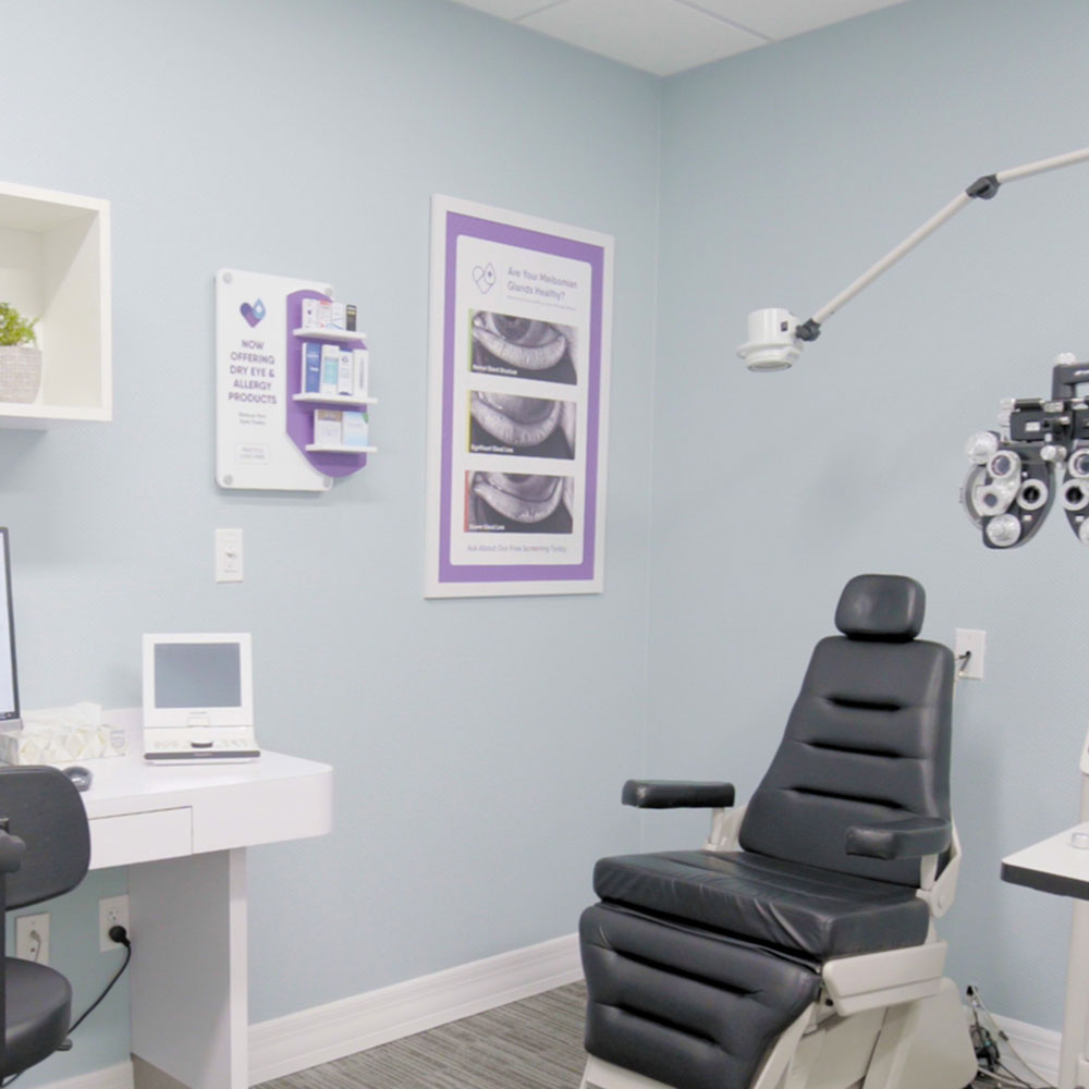 The exam room includes a black chair, an eye examination device by a white desk with a monitor, light blue walls adorned with informational posters and the DryEye Rescue PKG: Wall Mount Exam Room Display Conversation Starter (Small), nearby supply shelf, and overhead lamp for optimal interaction.