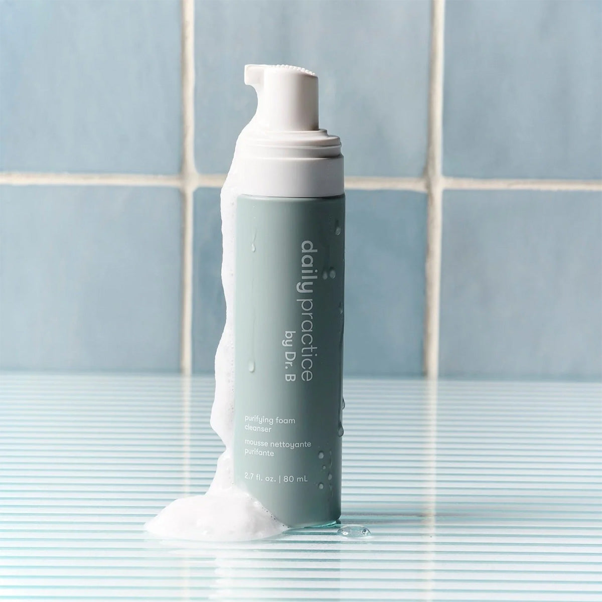 An 80mL bottle of Daily Practice Purifying Foam Cleanser, labeled Gentle & Hydrating for Eyes & Lashes, sits on a striped surface. Gentle foam overflows the top, enhancing the serene allure created by blue tiled walls in the background.