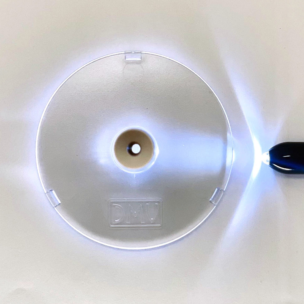 A clear DMV Scleral Contact Lens Inserter Stand with a vented scleral cup is on a white background. Illuminated by a right-sided light source, it creates a halo effect and displays the embossed DMV letters on its lower part.