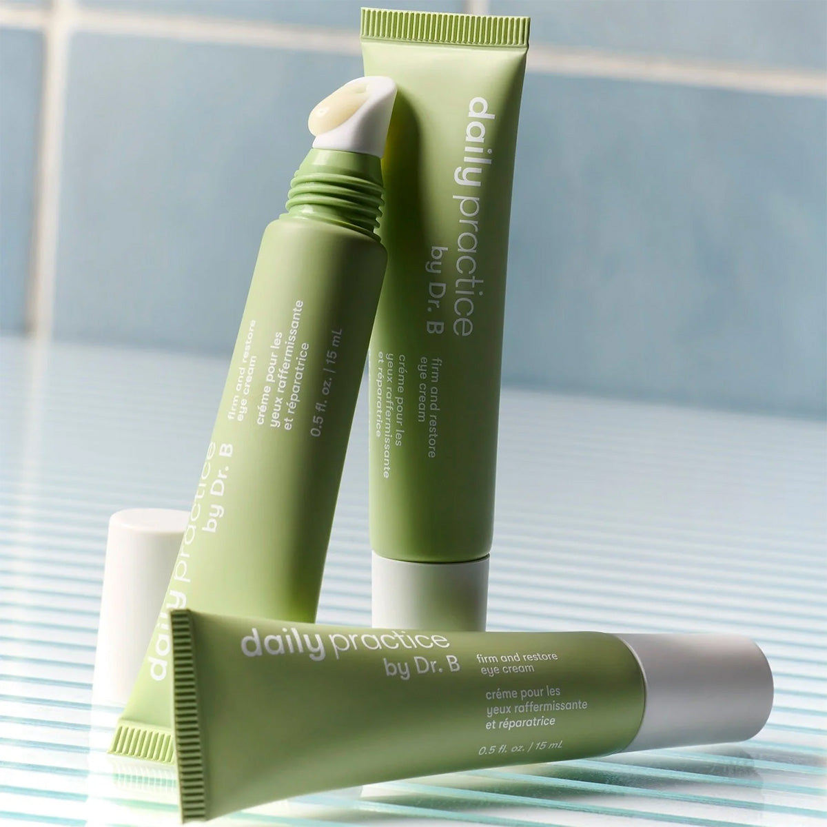 Three green tubes of Daily Practice Firm & Restore Eye Cream – Smooth, Hydrate & Lift (15mL) by Daily Practice are elegantly arranged on a striped surface. One uncapped tube reveals its creamy tip, showcasing its hydrating and firming effects on fine lines against a serene blue tiled backdrop.