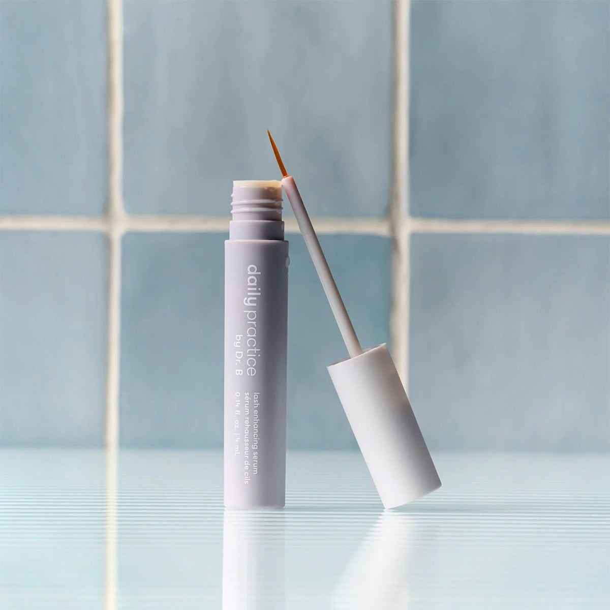 The Daily Practice Lash Enhancing Serum – Fuller, Longer, Healthier Lashes (4ml) stands upright on a reflective surface with its cap open, displaying a thin applicator. Blurred blue tiles in the background hint at its promise of natural eyelash growth.