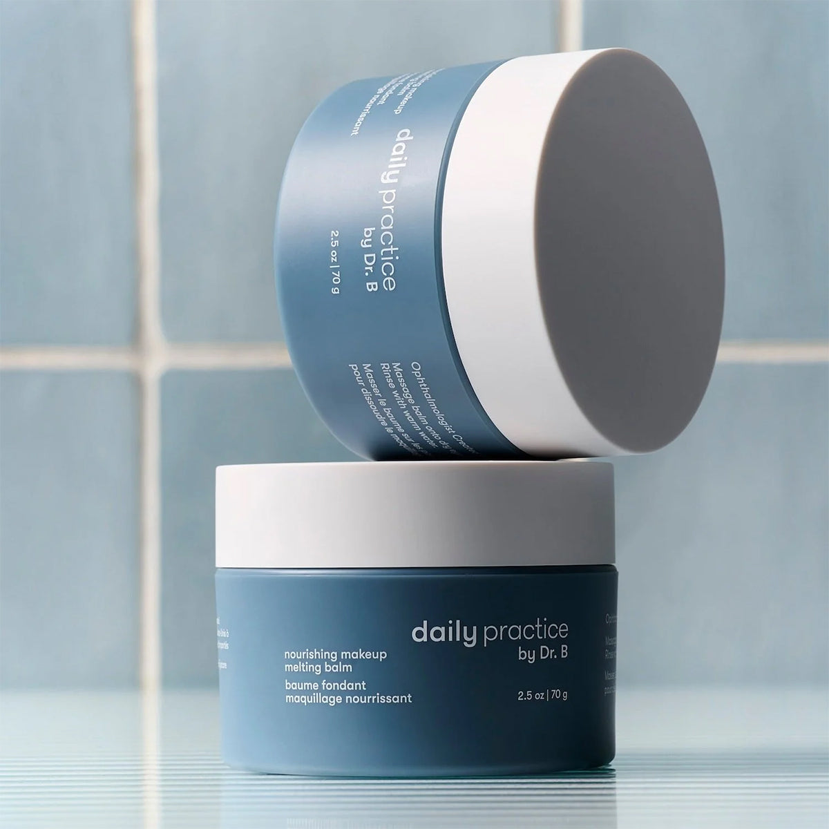 Two blue jars of Daily Practice Nourishing Makeup Melting Balm – Effortlessly Clean & Hydrate (70g) by Daily Practice, ophthalmologist-designed, are stacked on a reflective surface with a blue tiled wall background.