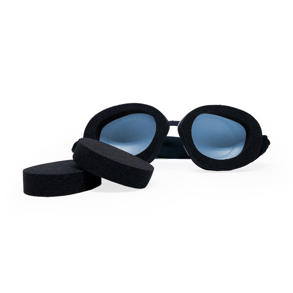 A pair of Eyeeco Tranquileyes Mini goggles by PRN, featuring blue-tinted lenses and memory foam for comfort, rests on a white surface. Nearby are two circular black foam pieces for padding or replacements—ideal for your Tranquileyes travel and sleep kit.